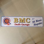 BMC 24 HOUR REPAIRS BANNER. On the left is the red, white and blue BMC rosette. Centre is BMC bold gold lettering. Dad's Garage lettering in red below. Right in blue lettering is For 24 Hour Repairs. White banner with blue edging.