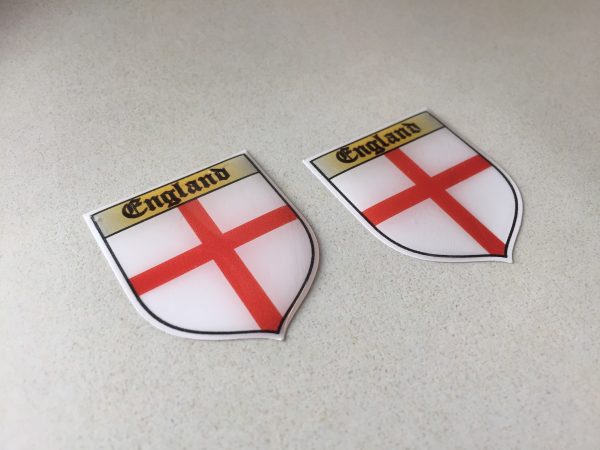 ENGLAND ST GEORGE SHIELD STICKERS DOMED RESIN GEL. A domed shield sticker. England in black Gothic text on a gold banner above a red cross on a white field.