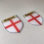 ENGLAND ST GEORGE SHIELD STICKERS DOMED RESIN GEL. A domed shield sticker. England in black Gothic text on a gold banner above a red cross on a white field.