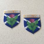 SCOTLAND EMBLEM SHIELD DOMED RESIN GEL STICKERS. Scotland in white lettering on a gold banner across the top of the shield. A thistle and a white saltire on a blue field. Domed sticker.