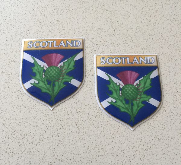 SCOTLAND EMBLEM SHIELD DOMED RESIN GEL STICKERS. Scotland in white lettering on a gold banner across the top of the shield. A thistle and a white saltire on a blue field. Domed sticker.