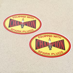 Champion in red lettering on a black dicky bow with a spark plug in the centre of a yellow oval sticker with a red border. Equipped with spark plugs lettering in red.