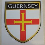 GUERNSEY FLAG SHIELD STICKER. Guernsey in white lettering on a black banner at the top of the shield. Below is a red cross with a gold cross within it on a white field.