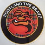 SCOTLAND THE BRAVE STICKER. Scotland The Brave in white lettering on a black outer circle. In the centre is a humorous face with red hair, beard and moustache on a red tartan background.
