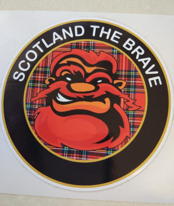 Scotland The Brave in white lettering on a black outer circle. In the centre is a humorous face with red hair, beard and moustache on a red tartan background.