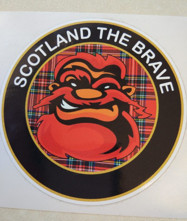 SCOTLAND THE BRAVE STICKER. Scotland The Brave in white lettering on a black outer circle. In the centre is a humorous face with red hair, beard and moustache on a red tartan background.