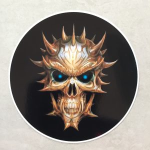 A multi horned skull in hues of brown and white with piercing vibrant blue eyes and sharp teeth. A circular sticker with a black background.