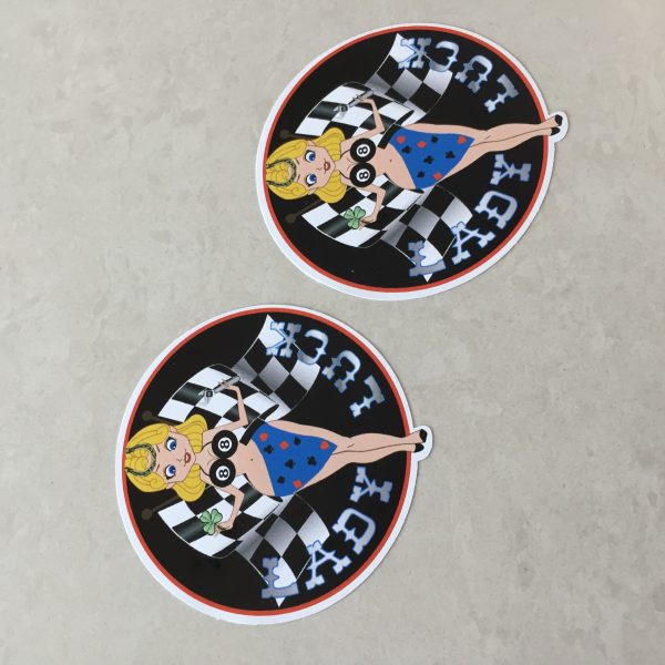 Lady Luck in silver lettering on a black background. A blonde pin up wearing a number 8 bikini top and four aces sarong with a horseshoe in her hair. She stands in front of crossed chequered flags while holding a tool and a clover leaf.