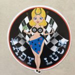 LADY LUCK STICKERS. Lady Luck in silver lettering on a black background. A blonde pin up wearing a number 8 bikini top and four aces sarong with a horseshoe in her hair. She stands in front of crossed chequered flags while holding a tool and a clover leaf.