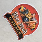 HOT ROD GARAGE STICKER. Hot Rod in white Gothic text, Garage in orange text on a black banner. A retro pin up in heels and a short dress leans over an orange hot rod with her gloved hands leaning on the wheel arch.