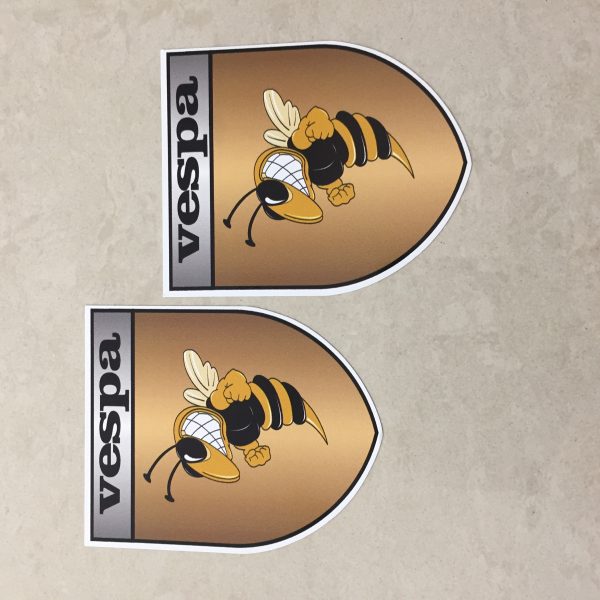 VESPA GOLD STICKERS. Vespa in black lettering on a silver banner at the top of the gold shield. A wasp displaying antennae, wings and white teeth is clenching its fists.