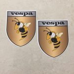 VESPA GOLD STICKERS. Vespa in black lettering on a silver banner at the top of the gold shield. A wasp displaying antennae, wings and white teeth is clenching its fists.