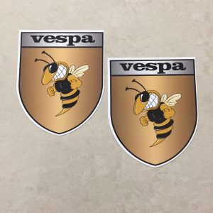 Vespa in black lettering on a silver banner at the top of the gold shield. A wasp displaying antennae, wings and white teeth is clenching its fists.