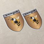 VESPA GOLD STICKERS. Vespa in black lettering on a silver banner at the top of the gold shield. A wasp displaying antennae, wings and white teeth is clenching its fists.