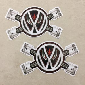 The VW logo overlays two crossed pistons. A pair of red evil eyes peer from behind the logo.