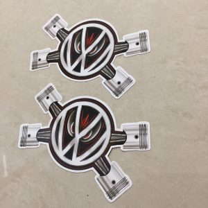 The VW logo overlays two crossed pistons. A pair of red evil eyes peer from behind the logo.