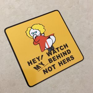 WATCH MY BEHIND STICKER. Hey! Watch My Behind Not Hers in black uppercase lettering on a yellow square sticker. A humorous female with yellow hair wearing a red dress and stockings is in the centre. She has her hand to her mouth while showing her behind.
