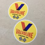 VALVOLINE RACING OIL STICKERS. Valvoline in red and Racing Oil in black uppercase lettering. Above is the letter V in red and blue. Below are two crossed chequered flags on a yellow circular sticker.