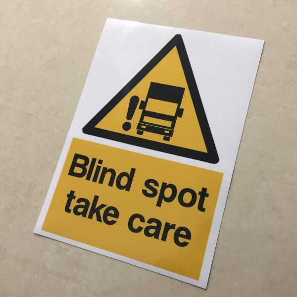 A black and yellow warning triangle featuring a lorry and an exclamation mark. Below in black lettering Blind spot take care within a yellow rectangle.