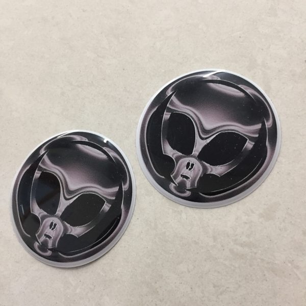 A grey/chrome effect domed circular sticker. A skull with large black eye sockets on a black background.