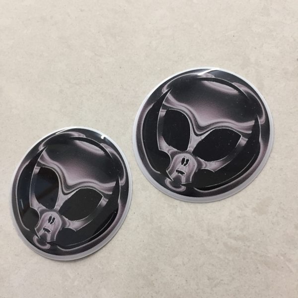 SKULL GEAR STICK CAP BADGE STICKERS. A grey/chrome effect domed circular sticker. A skull with large black eye sockets on a black background.