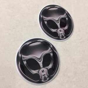 A grey/chrome effect domed circular sticker. A skull with large black eye sockets on a black background.