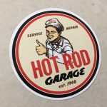 HOT ROD STICKER. A circular sticker with a black and red border. Hot Rod in red and Garage in black uppercase lettering. A man wearing a grey cap and jacket is winking and giving the thumbs up. Additional lettering Service, Repair, Est. 1948.