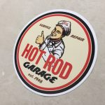HOT ROD STICKER. A circular sticker with a black and red border. Hot Rod in red and Garage in black uppercase lettering. A man wearing a grey cap and jacket is winking and giving the thumbs up. Additional lettering Service, Repair, Est. 1948.