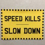 SPEED KILLS BIN STICKER. Speed Kills Slow Down in black uppercase lettering on a yellow sticker bordered with black squares equidistance apart.