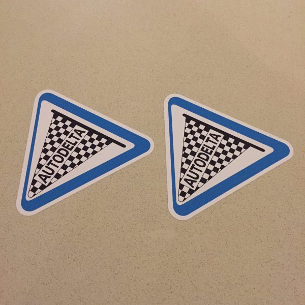 AUTODELTA STICKERS. Autodelta in black uppercase lettering on a black and white chequered flag and pole. A white triangular sticker with a blue border.