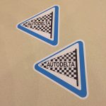 AUTODELTA STICKERS. Autodelta in black uppercase lettering on a black and white chequered flag and pole. A white triangular sticker with a blue border.