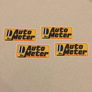 AUTO METER STICKERS. A gauge in black and white. Auto Meter in black. Competition Instruments in red lettering on a yellow sticker with a red border.
