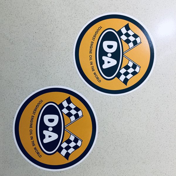 DA ENGINE OIL STICKERS. An orange circular sticker. Two crossed chequered flags sit above D A in white letters on a black oval. Toughest Engine Oil In The World in black lettering below.