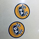 DA ENGINE OIL STICKERS. An orange circular sticker. Two crossed chequered flags sit above D A in white letters on a black oval. Toughest Engine Oil In The World in black lettering below.