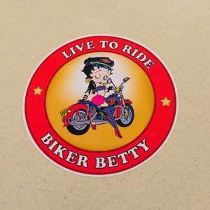 LIVE TO RIDE BETTY BOOP STICKER. Live To Ride Biker Betty in white lettering surrounds a red circular sticker. Centre is a cartoon character Betty Boop astride a motorcycle, registration plate BOOP. She is wearing skimpy colourful clothing, stockings and suspenders.