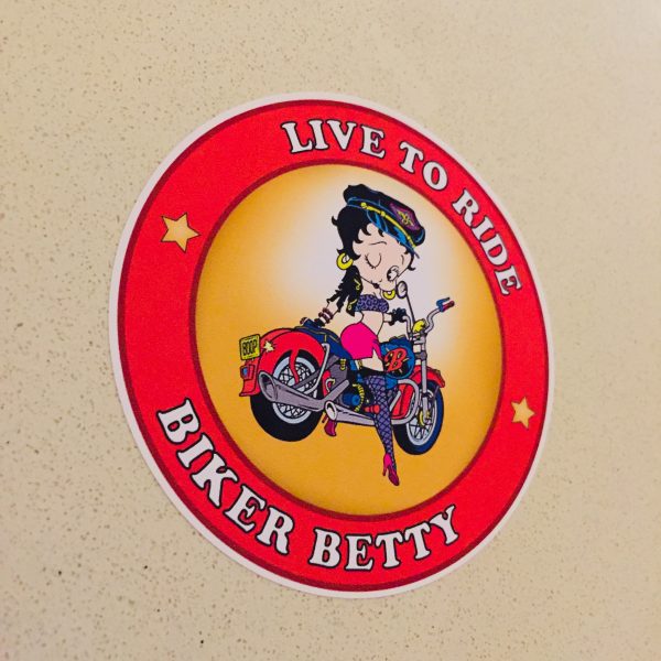 LIVE TO RIDE BETTY BOOP STICKER. Live To Ride Biker Betty in white lettering surrounds a red circular sticker. Centre is a cartoon character Betty Boop astride a motorcycle, registration plate BOOP. She is wearing skimpy colourful clothing, stockings and suspenders.