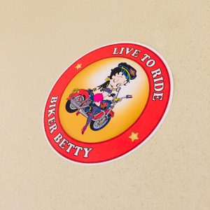 Live To Ride Biker Betty in white lettering surrounds a red circular sticker. Centre is a cartoon character Betty Boop astride a motorcycle, registration plate BOOP. She is wearing skimpy colourful clothing, stockings and suspenders.