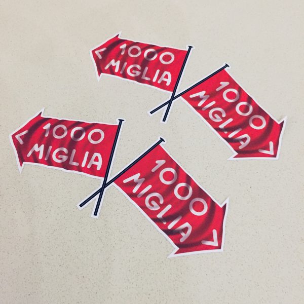Two crossed flags. 1000 Miglia in white lettering on a red direction of travel arrow with a white border.