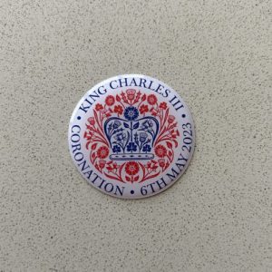 KING CHARLES CORONATION DOMED RESIN GEL STICKER. King Charles lll Coronation 6th May 2023 in blue lettering surrounds this white domed sticker. In the centre is a blue crown made up of roses, thistles, daffodils and shamrocks. These flowers, in red, surround the crown.