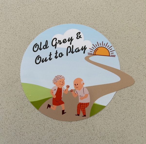 OLD GREY AND OUT TO PLAY STICKER. Old, grey and out to play in black text on a circular sticker. An elderly couple having fun on a winding path surrounded by green fields, blue sky and sunshine.