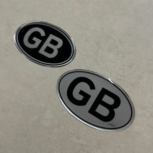 GB STICKER DOMED RESIN GEL. GB in uppercase lettering on an oval sticker. Black letters on chrome or chrome letters on a black background.